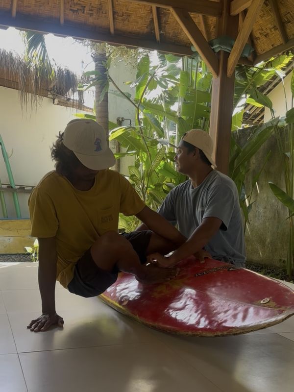 Two surfers waxing a board in the shade of a tropical camp