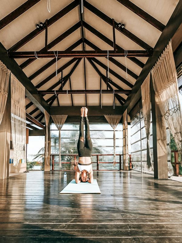 A person in a headstand in a tall, timber-roofed practice hall open to the trees