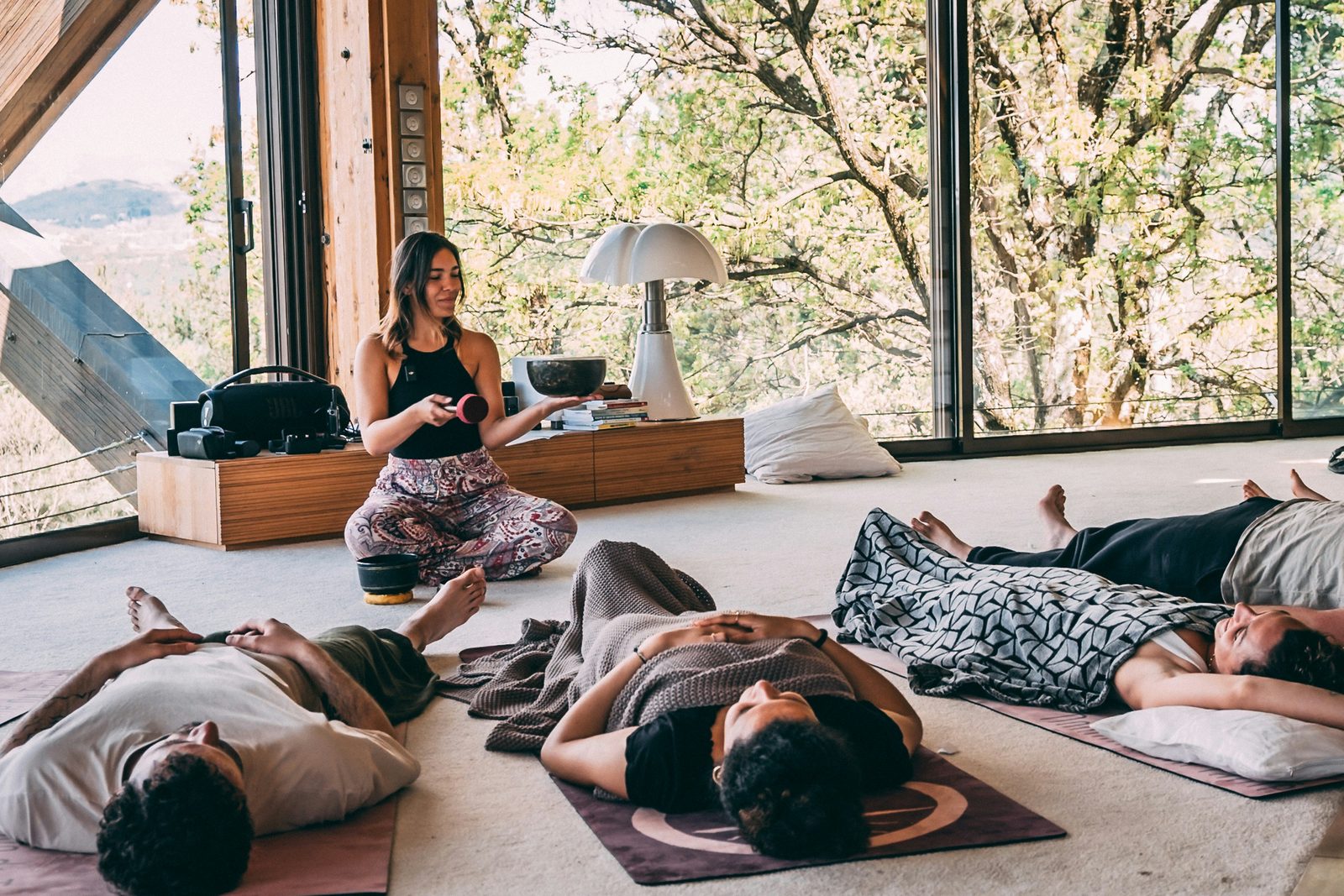 A retreat group resting on their mats while their teacher plays a singing bowl, in a bright room among the trees