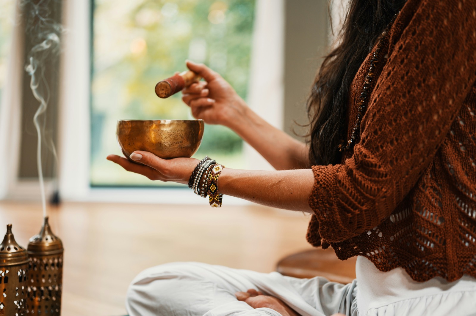 A singing bowl held in two hands beside rising incense smoke, in a sunlit room